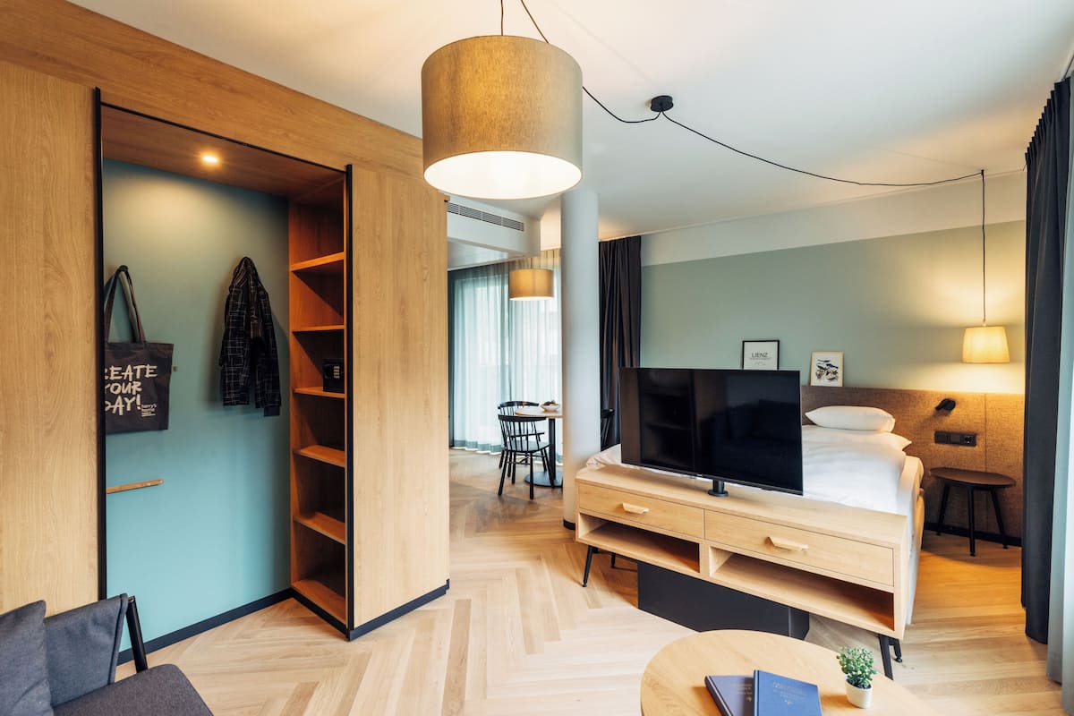 A modern hotel room with a wooden wardrobe, a TV, a bed, a desk, and a dining area.