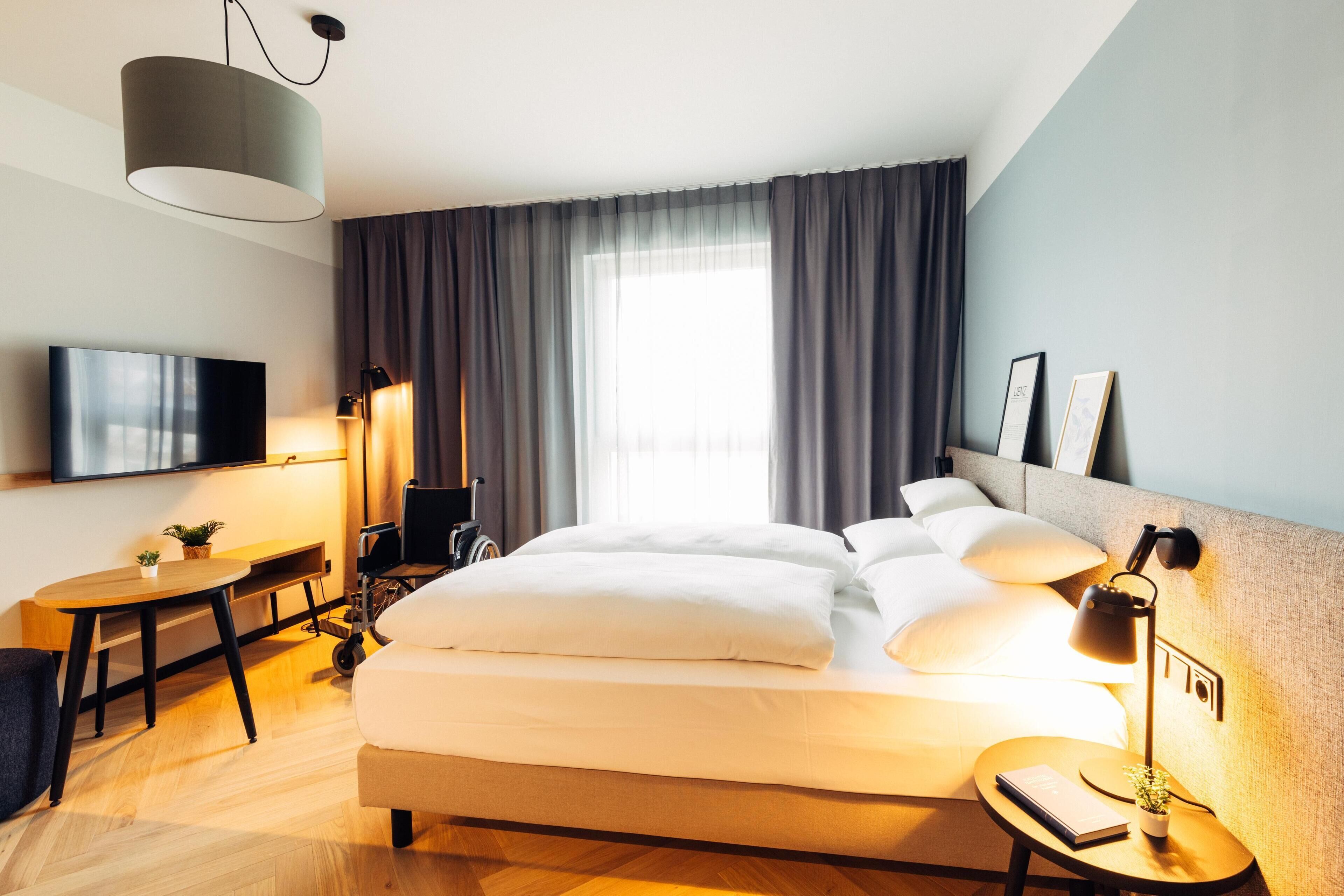 Una habitación de hotel moderna con una cama grande, mesita de noche, lámpara y televisor en la pared.