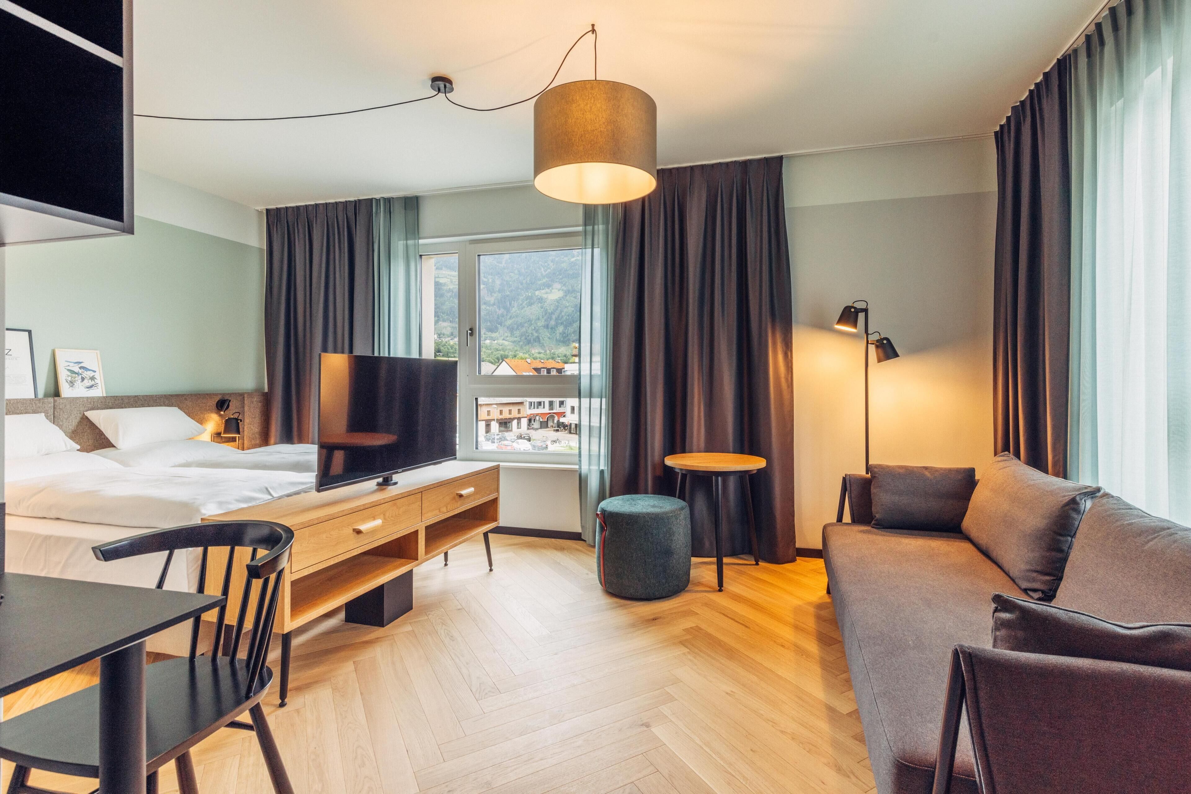 Quarto de hotel moderno com cama, escrivaninha, sofá, televisão e janela com cortinas.