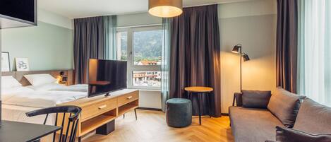 A modern hotel room with a bed, a desk, a sofa, a TV, and a window with curtains.