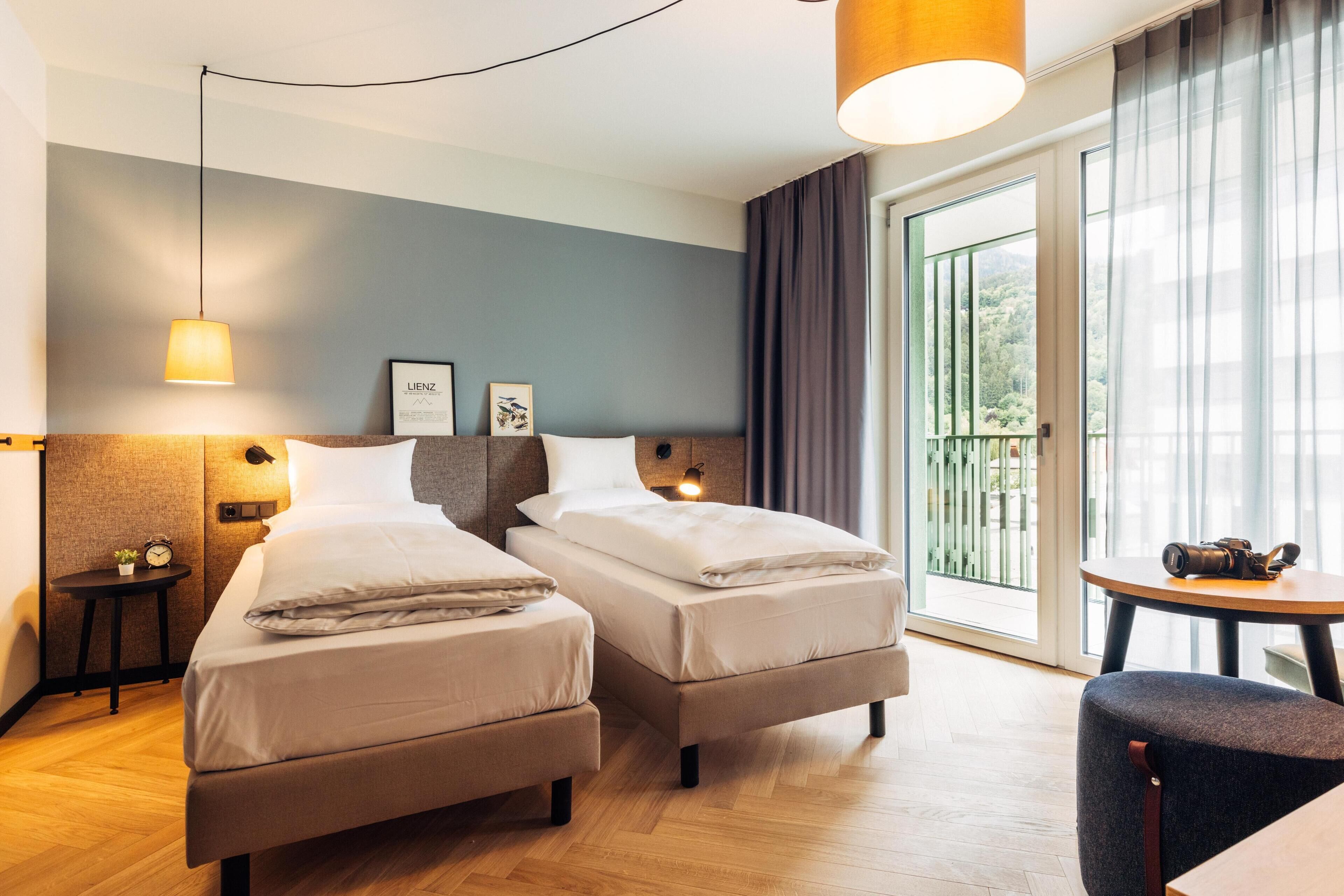 Habitación de hotel con dos camas, una mesita, una silla, una lámpara y vista al exterior.