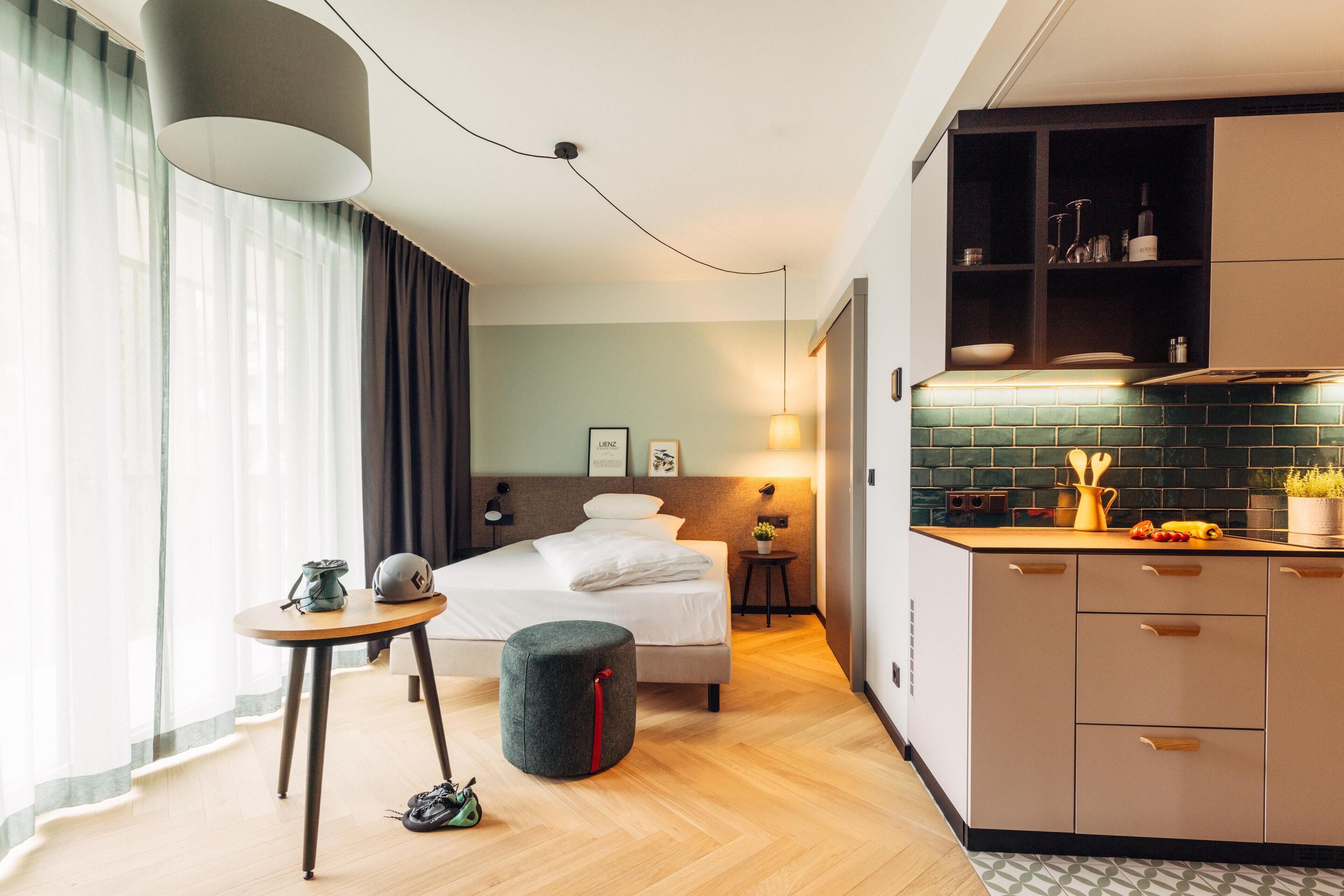 A modern hotel room with a bed, bedside table, and a small stool. There is a kitchenette with a stove, sink, and cabinets.