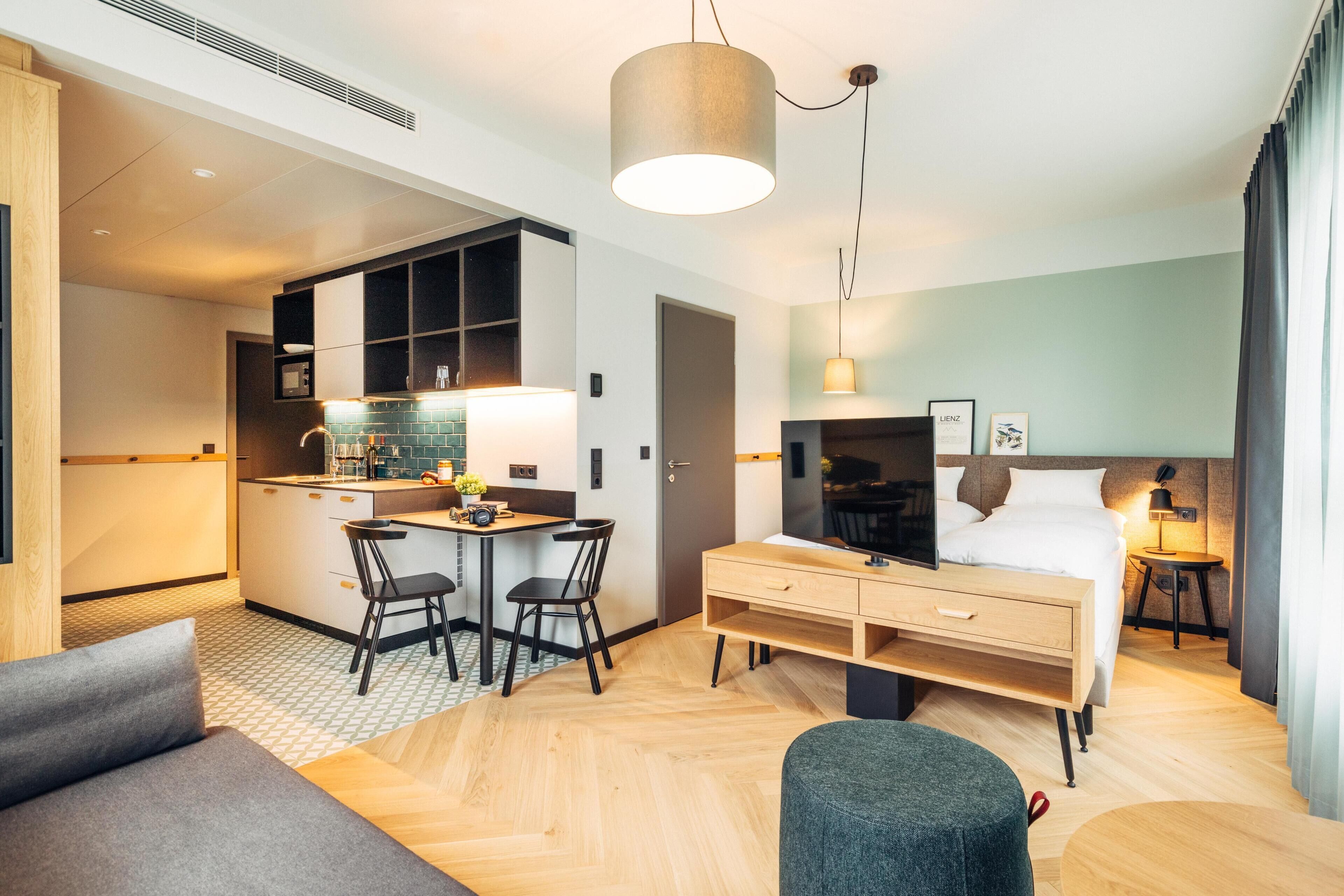 Habitación de hotel moderna con kitchenette, zona de comedor, cama y un escritorio pequeño.