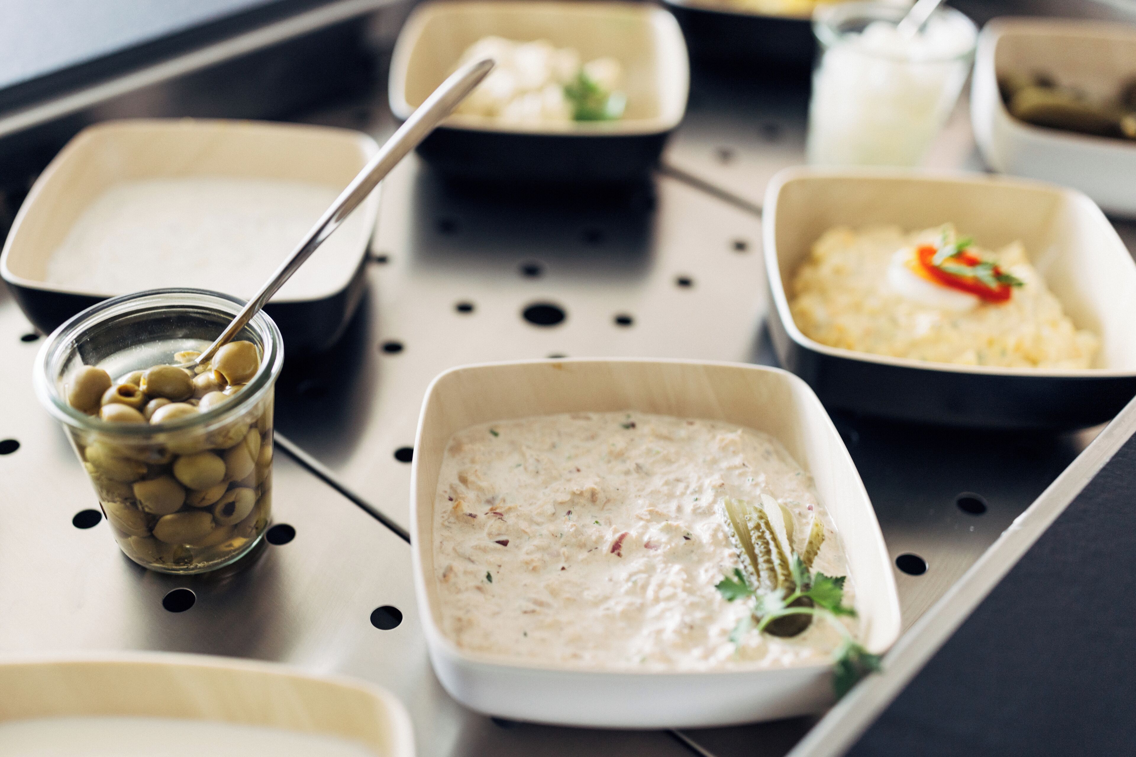 Egy tálca, melyen különféle ételek találhatók, többek között egy krémes étel, egy tál olívabogyó és egy kanállal ellátott edény.