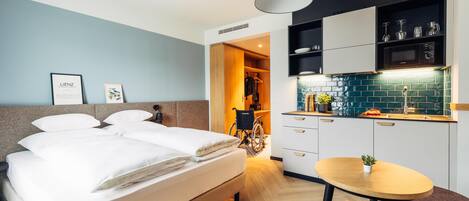 A modern hotel room with a bed, a small dining table, a kitchenette, and a wheelchair-accessible bathroom.