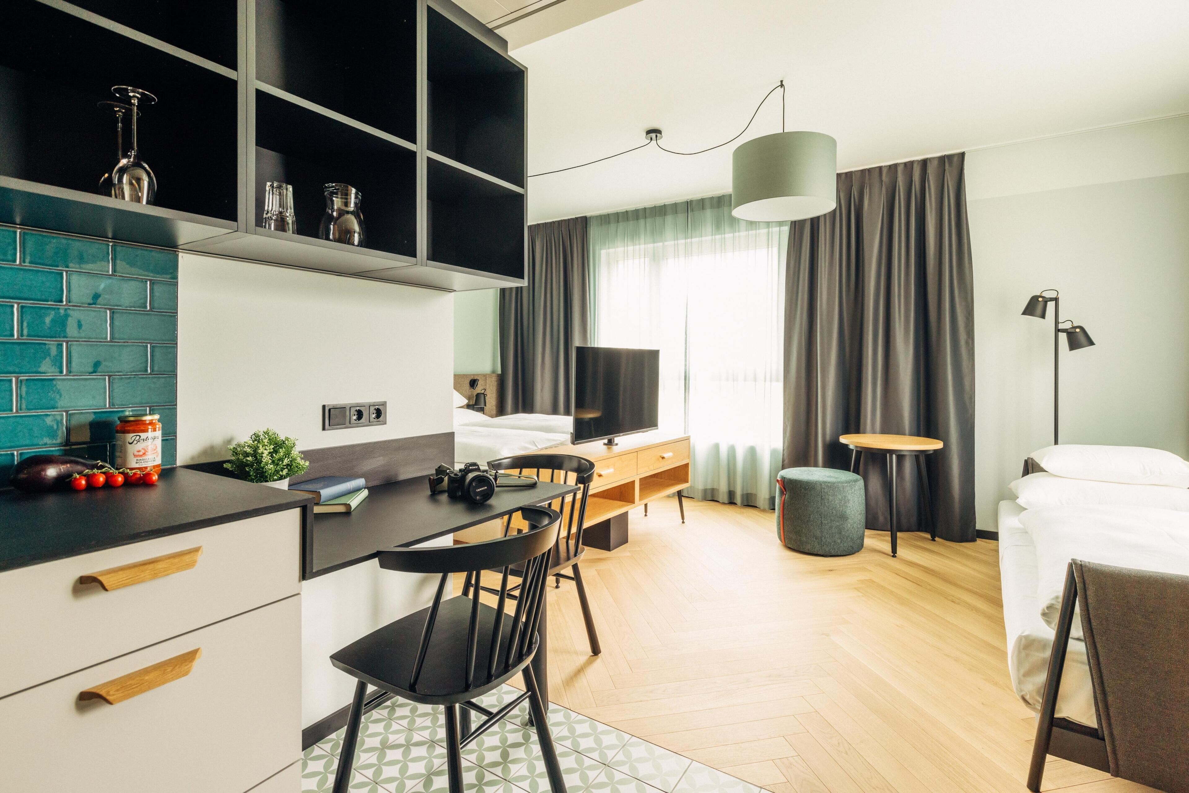 A modern hotel room with a kitchen area, a dining table, and a bed.