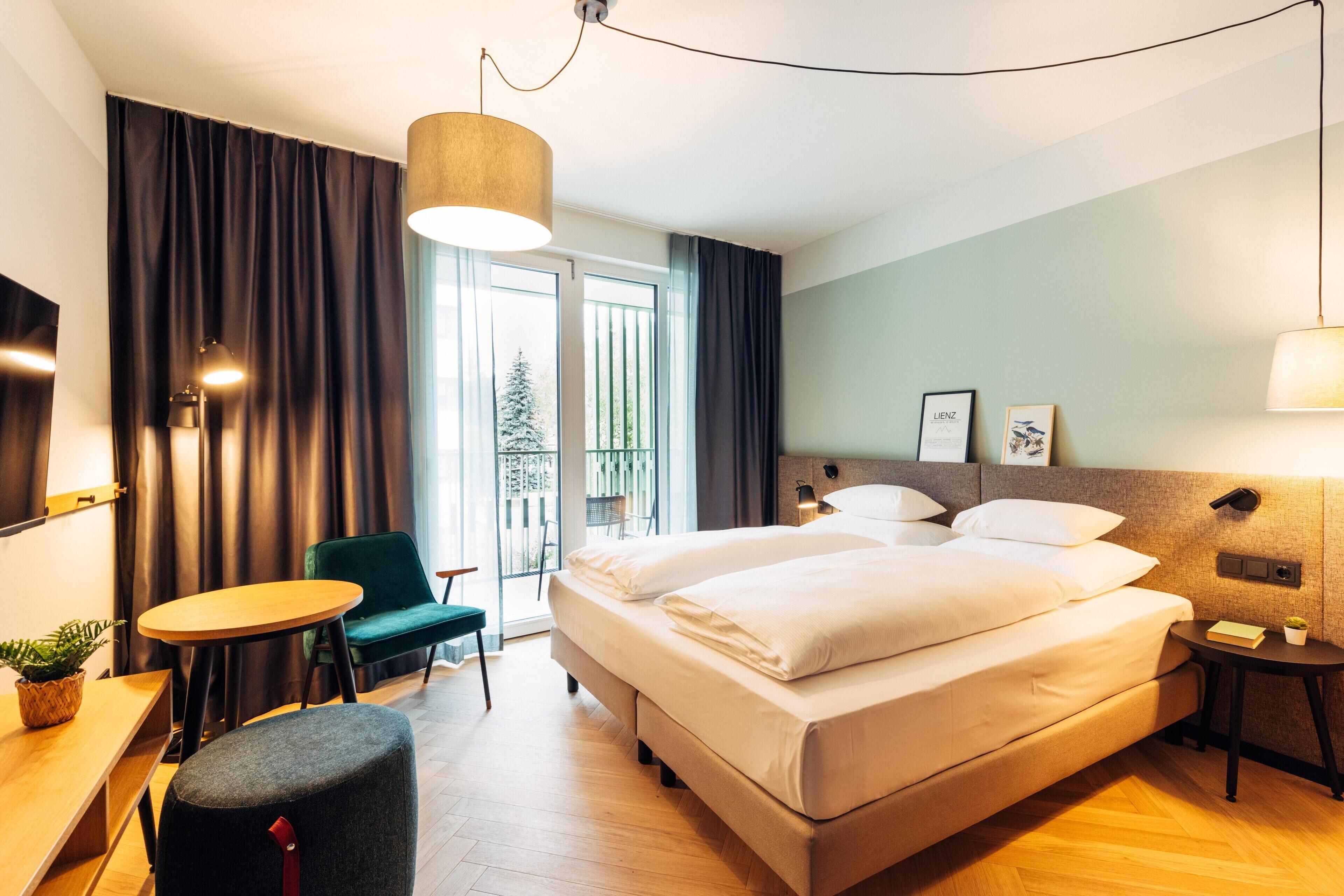 A hotel room with a large bed, a desk, a chair, a small table, and a balcony with a view of trees.