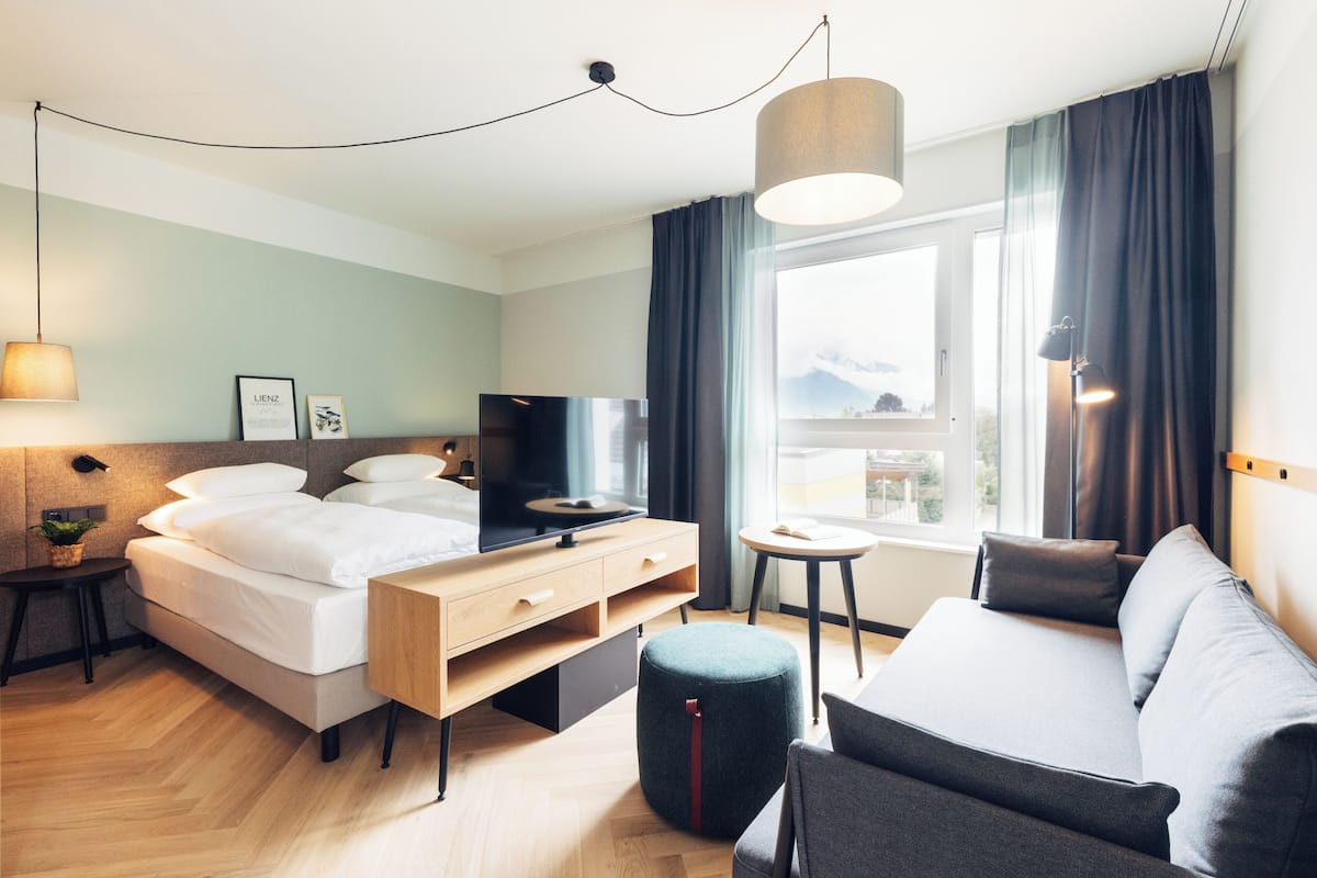 A modern hotel room with a bed, a sofa, a TV, a small table, and a window with a view of mountains.