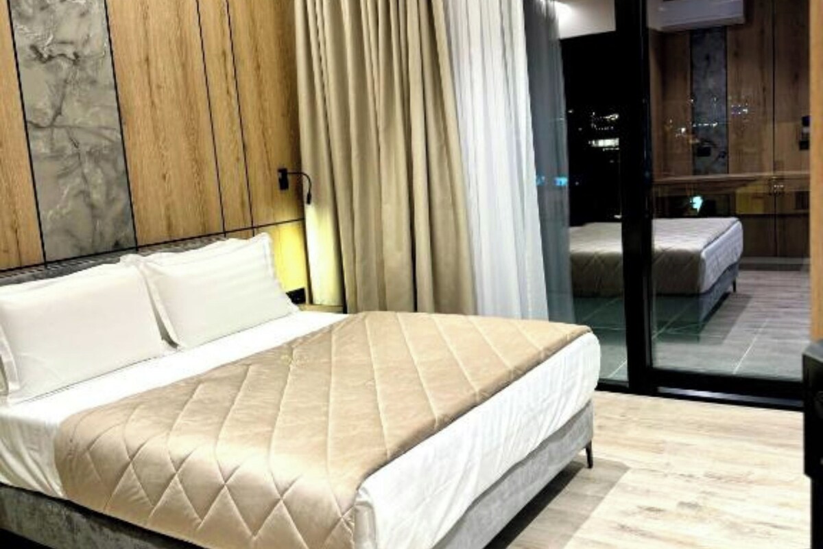 A modern bedroom with a large bed, wooden panel wall, and a view through a glass door.