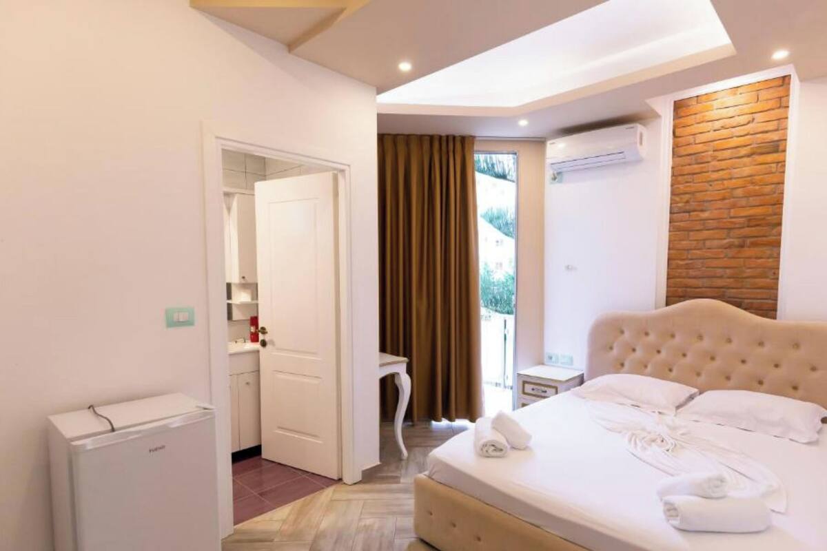 A modern hotel room with a large bed, a compact kitchenette, and a view of the outdoors through a window.