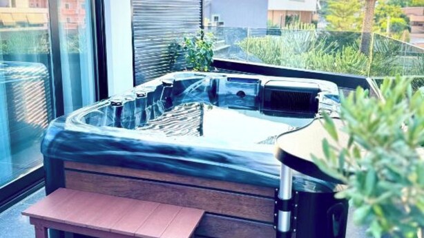 A hot tub with a wooden bench in front, surrounded by glass walls and plants. - Hotel Joni Premium  (Ksamil)