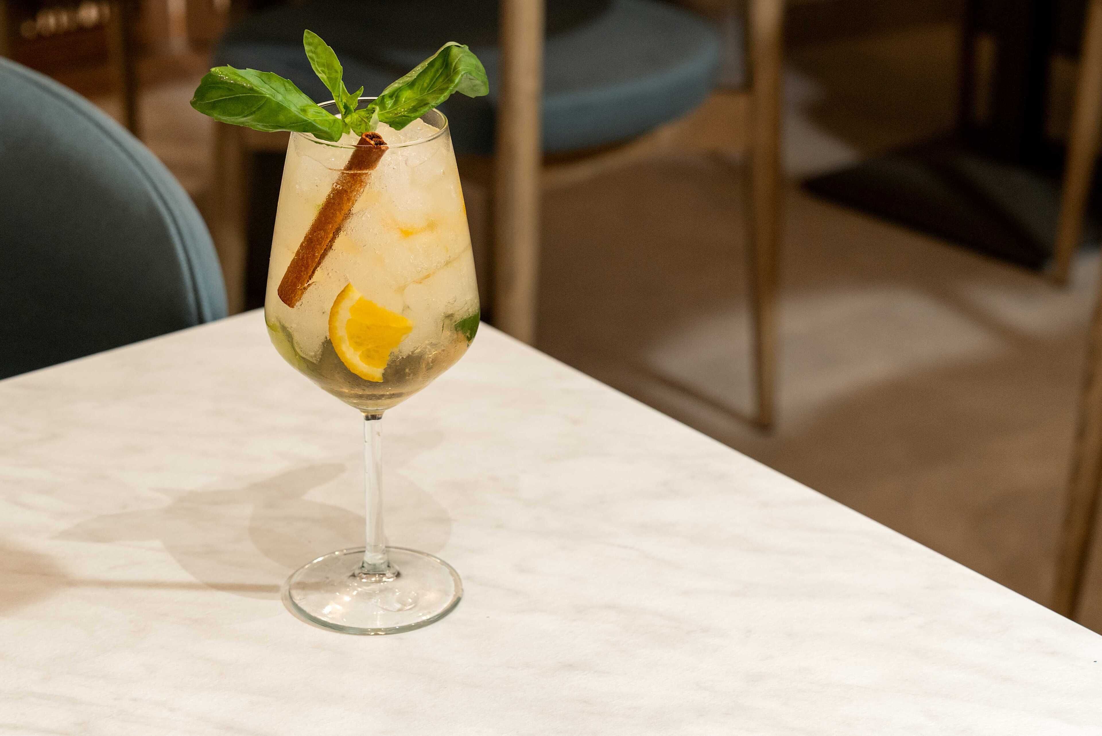 A cocktail glass with ice, lemon, mint leaves, and a cinnamon stick on a marble table.