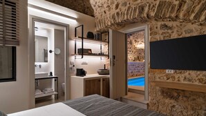 A bedroom with a stone wall, a TV mounted on the wall, a bed, a bedside table, a bathroom with a sink and toilet, and a view of a pool. - YeeOn Boutique Hotel, Gythio - Historic Hotels of Europe (East Mani)