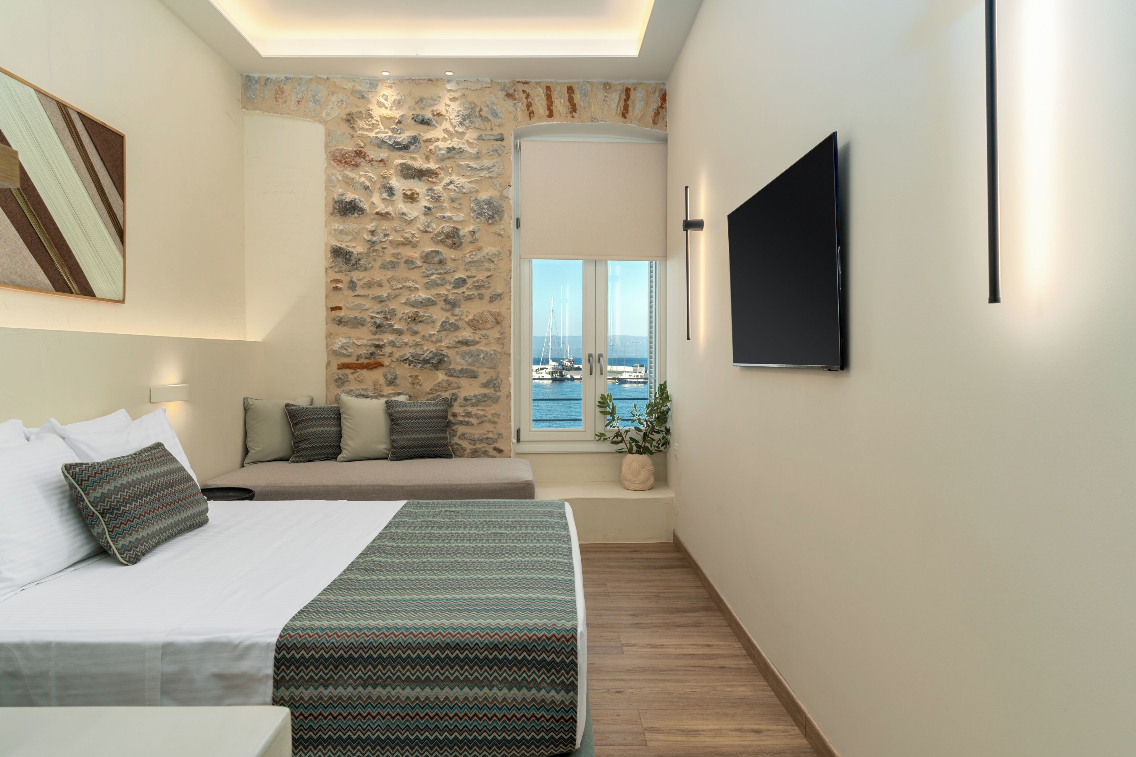 Una habitación de hotel moderna con una cama grande, una zona de estar con vistas al mar y un televisor de pantalla plana montado en la pared.