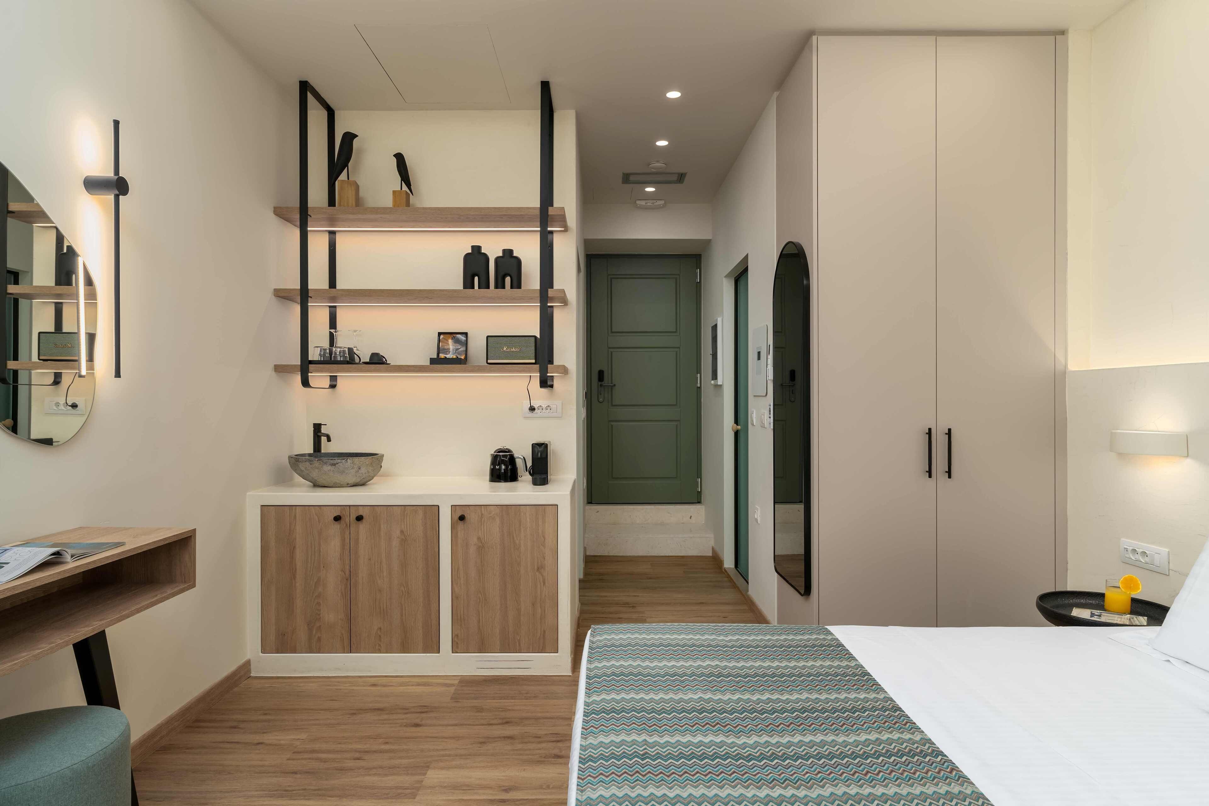 Een moderne hotelkamer met een bed, een bureau, een kaptafel met spiegel, planken met decoratieve objecten, en een groene deur op de achtergrond.