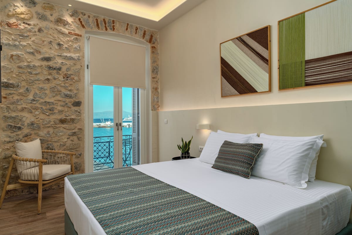 A bedroom with a stone wall, a bed with white linens, a view of the sea through a balcony door, and a potted plant on the nightstand.