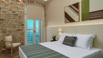 Un dormitorio con pared de piedra, una cama con ropa blanca, vista al mar a través de una puerta de balcón y una planta en maceta sobre la mesita de noche.