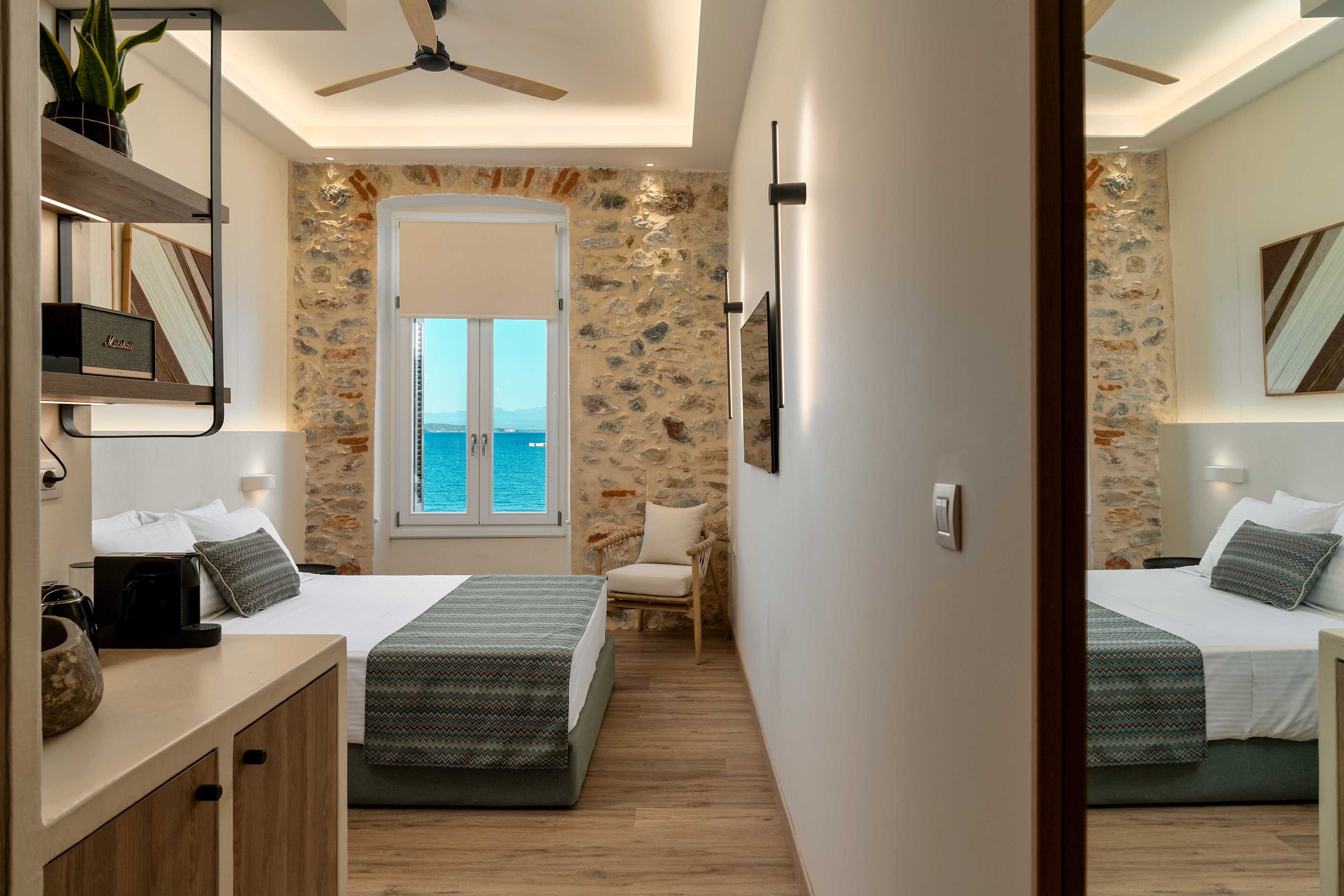 A hotel room with two beds, a desk, a chair, a ceiling fan, and a view of the sea through a large window.