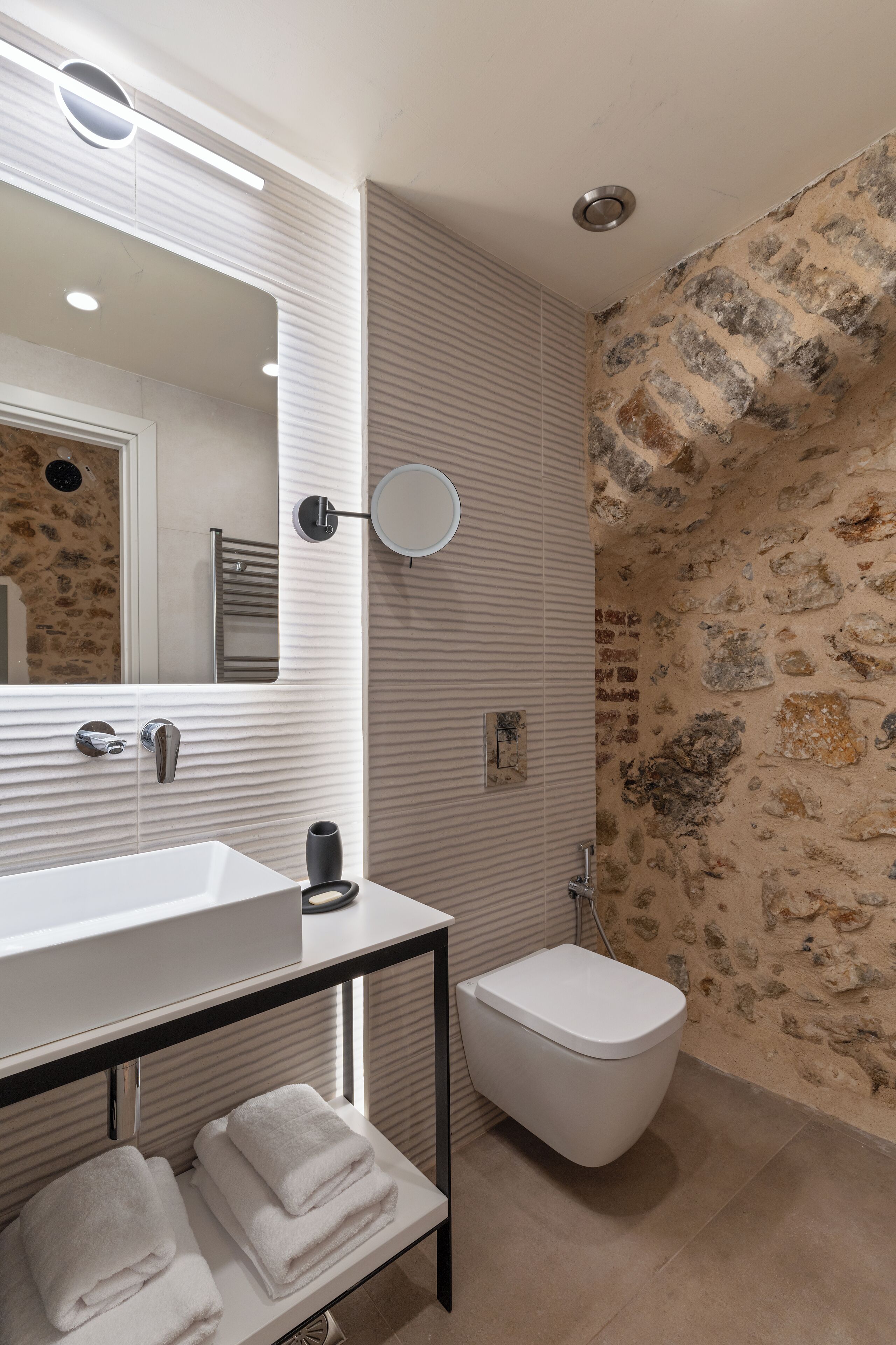 A bathroom with a white sink, a mirror, a towel rack, a toilet, and a stone wall.