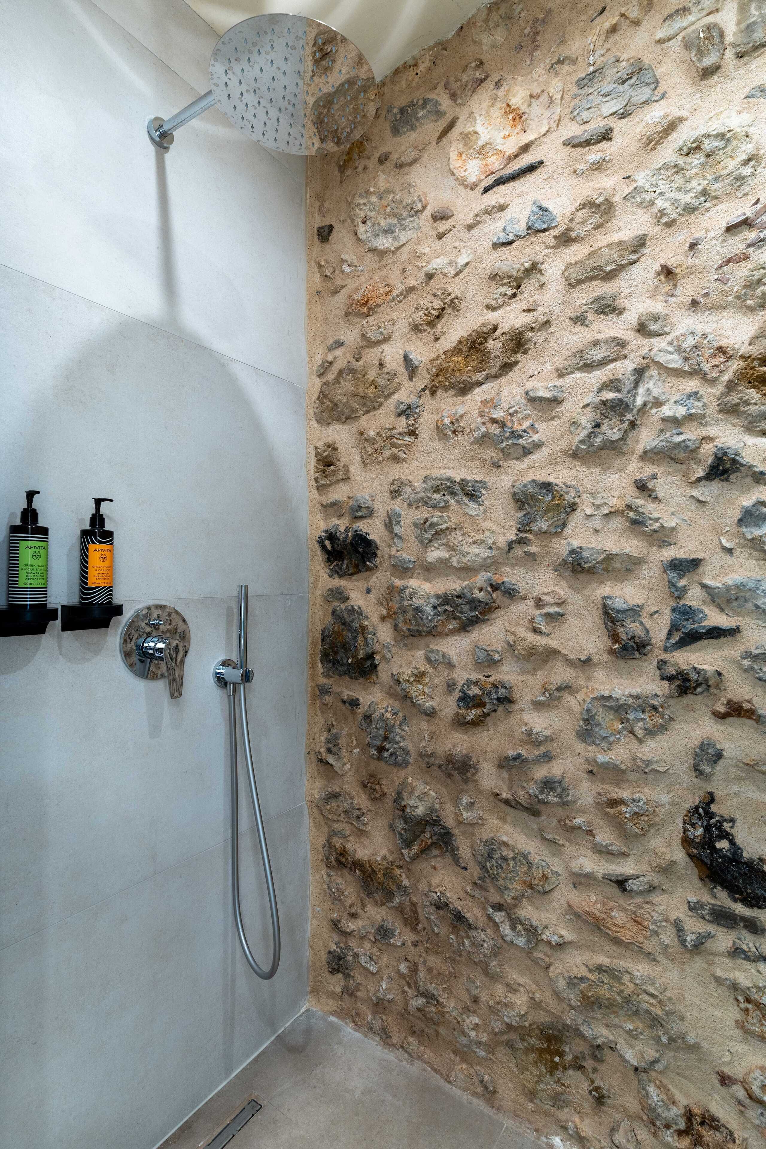 Ducha con pared de piedra, cabezal de ducha de mano y dos botellas de champú en un estante.