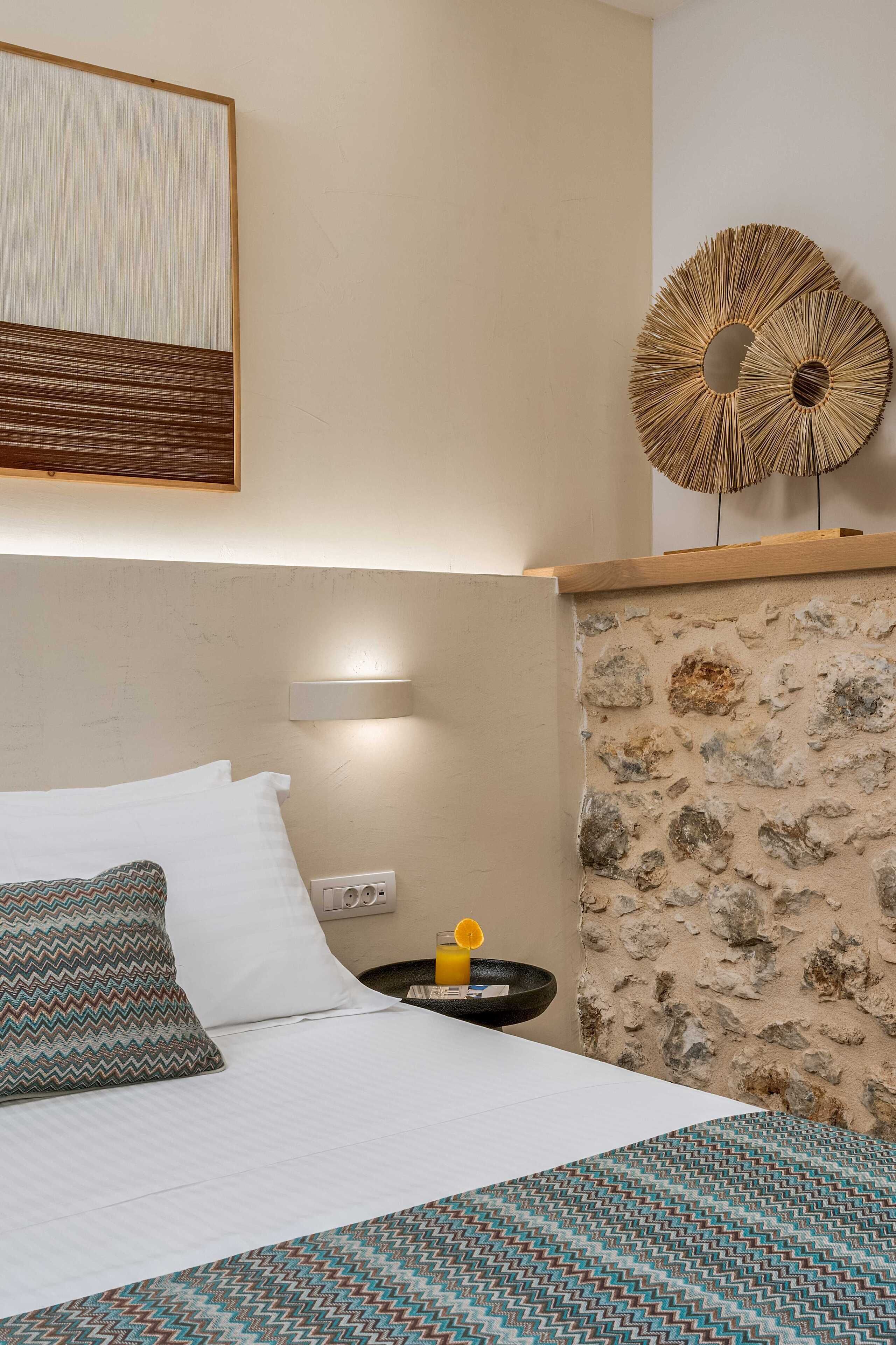 Una habitación de hotel con una cama, un espejo fijado a la pared, un adorno mural y una mesita de noche con una lámpara, y una pared con textura de piedra.