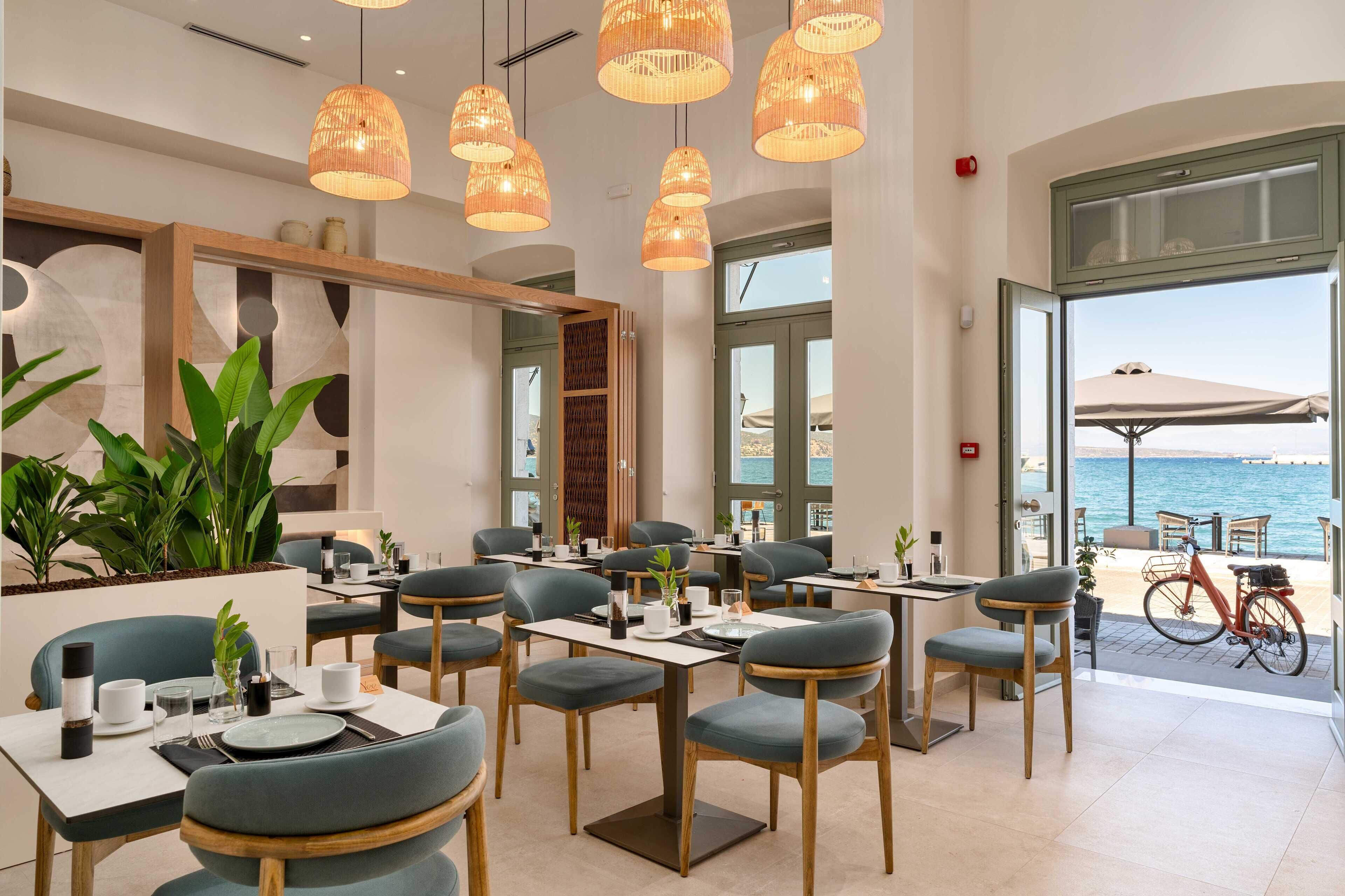 A modern dining area with tables and chairs, large windows, and a view of the sea.
