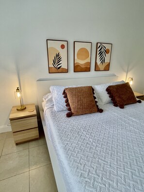 A neatly made bed with white bedding and two brown pillows, flanked by a nightstand with a lamp and a drawer. Three framed artworks are hung on the wall above the bed.