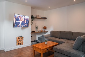 A living room with a grey sofa, a wooden coffee table, a TV mounted on the wall, and a fireplace with logs.