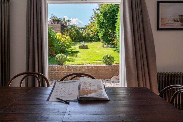 A wooden table with a menu and a pen, a window with curtains, and a view of a garden with a brick wall and greenery.