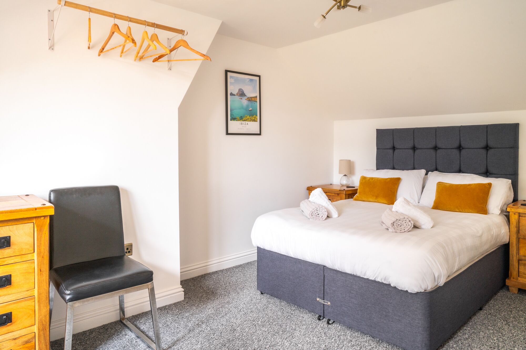 A bedroom with a bed, a desk, a chair, a hanging clothes rack, and a framed picture on the wall.
