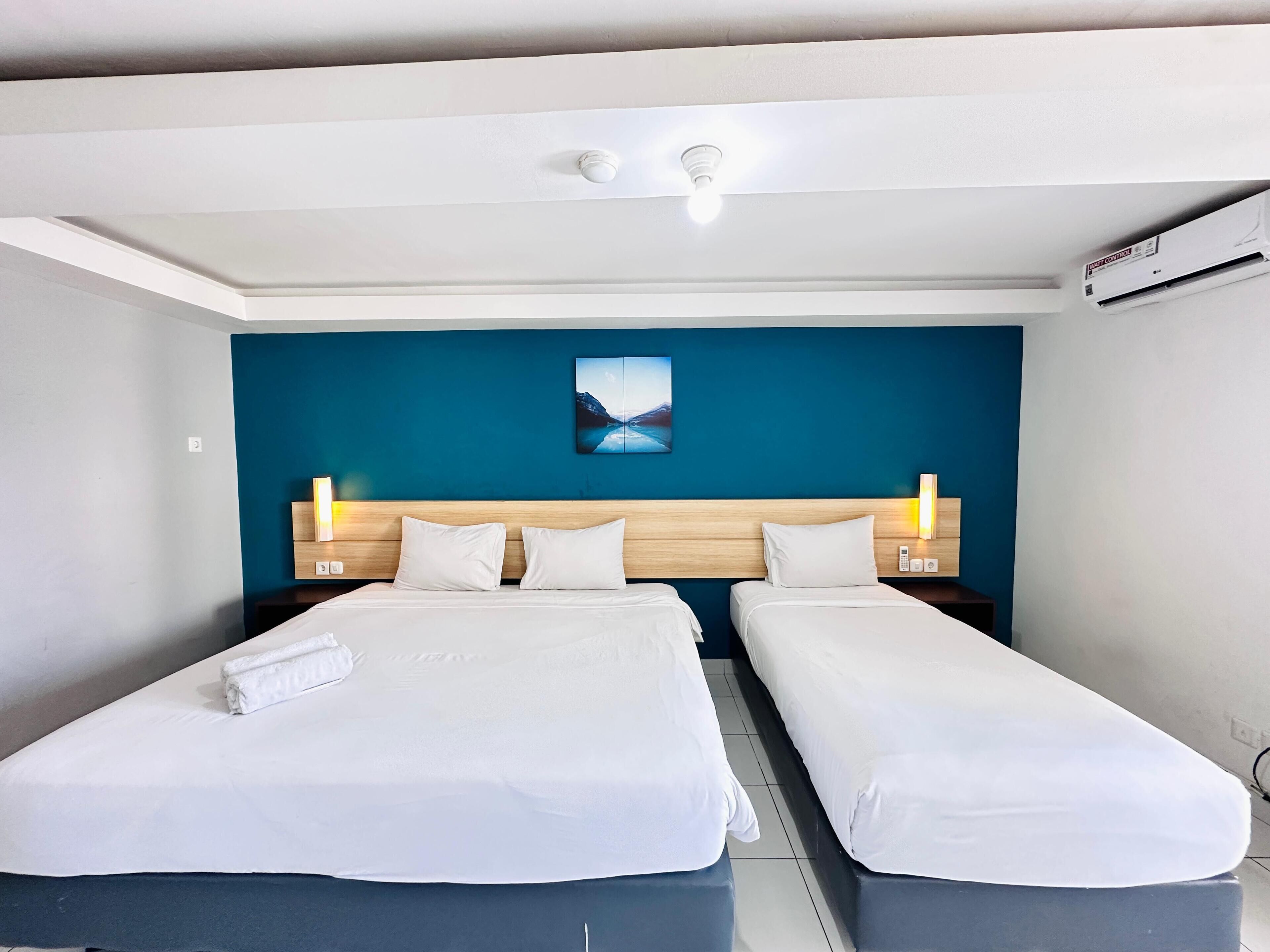 Habitación de hotel con dos camas, cabecero de madera, pared azul decorativa, un cuadro y un equipo de aire acondicionado.