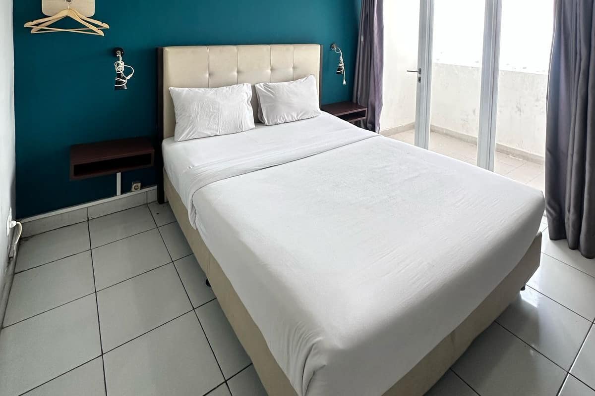 A bedroom with a large bed, a wooden headboard, a nightstand, a wall-mounted light, and a view of the outside through a window.