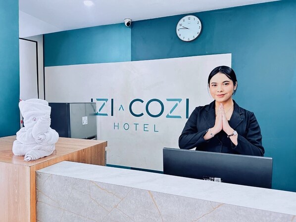 A hotel reception desk with a clock, a sign reading "ZI A COZI HOTEL", and a stack of white towels.