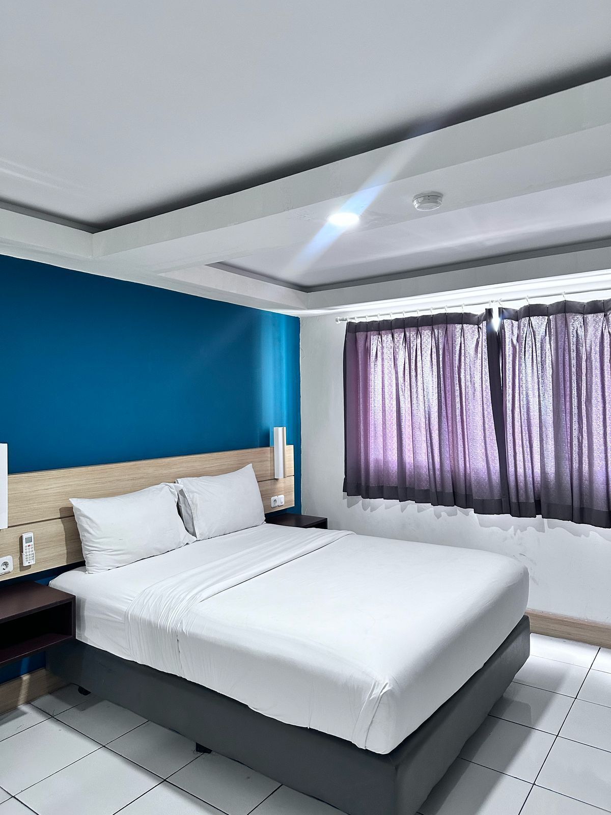 Ein modernes Hotelzimmer mit einem großen Bett, einem Nachttisch, einem Beistelltisch und einem Fenster mit violetten Vorhängen.