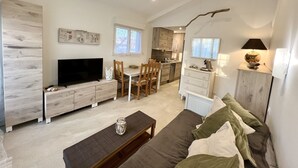 En moderne stue med et fladskærms-tv, træmøbler, et spiseområde med stole og et køkken i baggrunden.