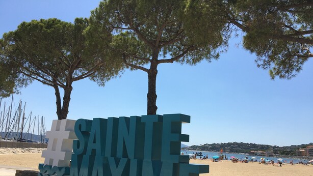 Et stort '#SAINTE MAXIME'-skilt i et kystområde med en strand og sejlbåde i baggrunden.