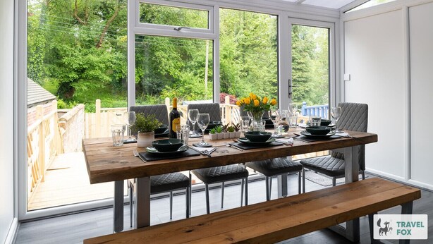 Un invernadero con una mesa de madera dispuesta para comer, rodeada de sillas y grandes ventanales con vistas a la vegetación.