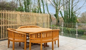 Ein Holztisch mit Stühlen steht auf einer Terrasse, im Hintergrund eine Holzlattenveranda und Grünpflanzen.