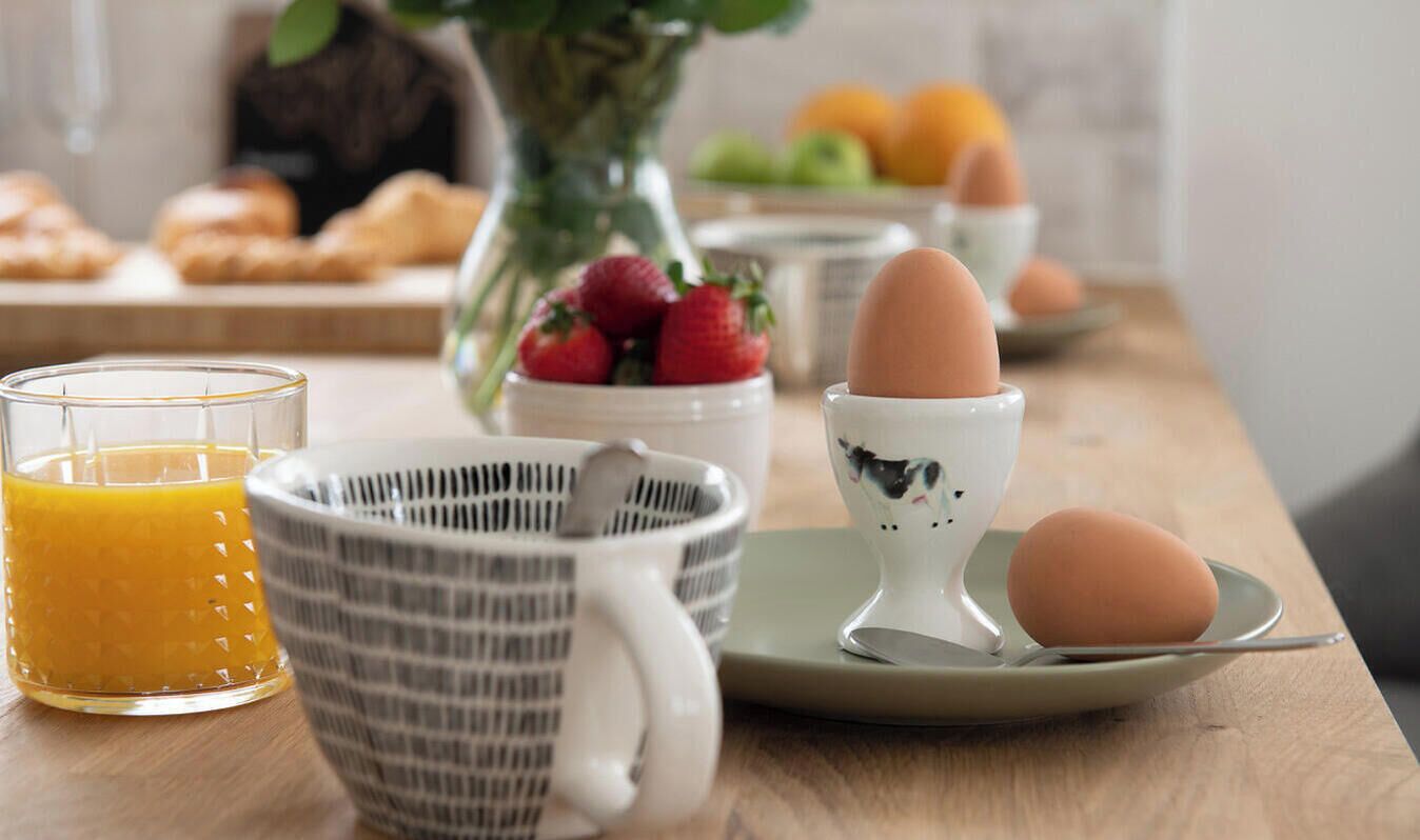 Colazione con un bicchiere di succo d'arancia, una tazza a righe, una ciotola di fragole, un portauovo in ceramica e un piatto con un uovo.
