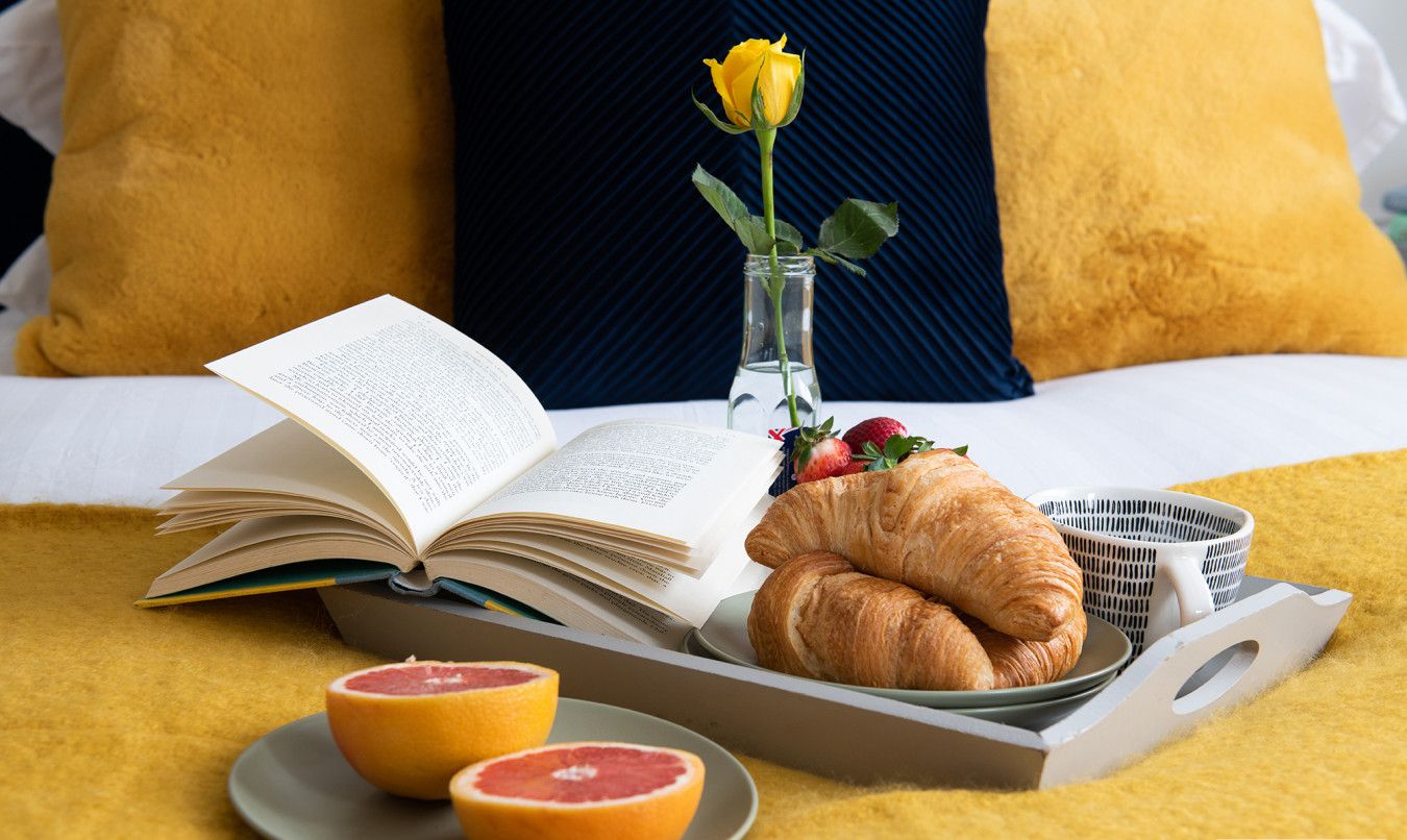 Ein Frühstücksarrangement mit einem offenen Buch, zwei Croissants, einem Glas Orangensaft, einer Rose in einer Vase und einem Korb mit einer Erdbeere.