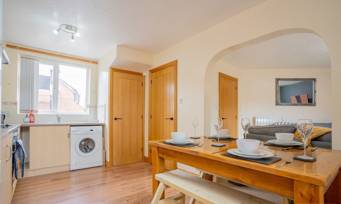 A compact kitchen with a washing machine, wooden cabinets, and a dining area with a bench.