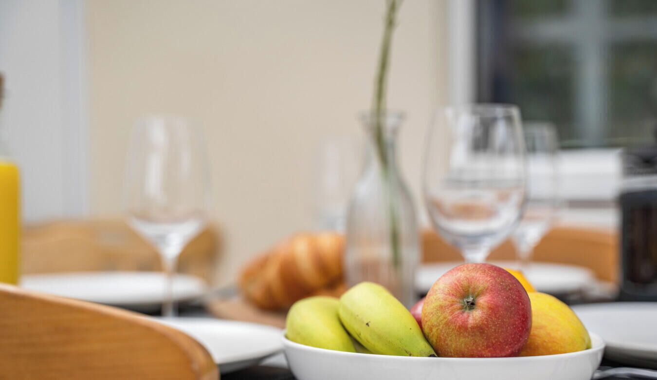 Una mesa de comedor con un bol de fruta y un croissant, junto con copas de vino.