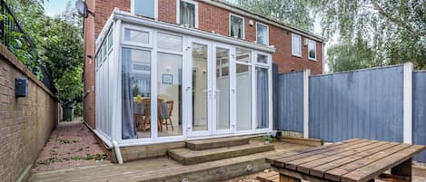 A conservatory with a wooden deck, a table, and a brick house in the background.
