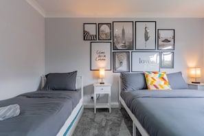 A twin bedroom with two beds, a nightstand with a lamp, and a wall with framed pictures. - Jodi's Bungalow, Parking & Large Garden (Ruddington)