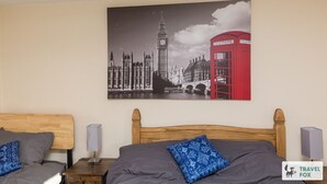 Une chambre d’hôtel avec deux lits, un grand tableau représentant un paysage urbain accroché au mur et une cabine téléphonique rouge.