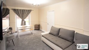 Une pièce avec un canapé gris, une petite table, une chaise, un téléviseur et une fenêtre avec des rideaux.