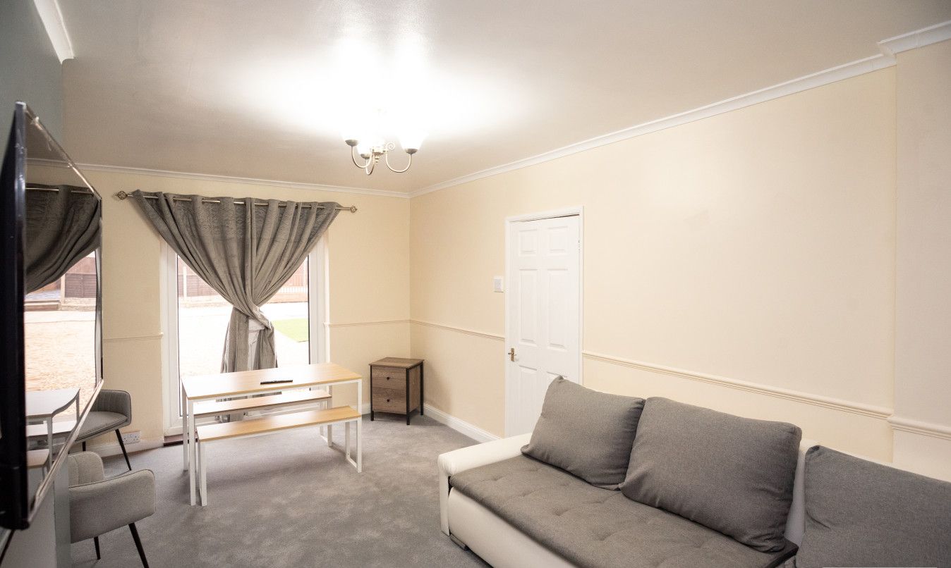 A room with a grey sofa, a small table, a chair, a TV, and a window with curtains.