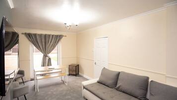 A room with a grey sofa, a small table, a chair, a TV, and a window with curtains.