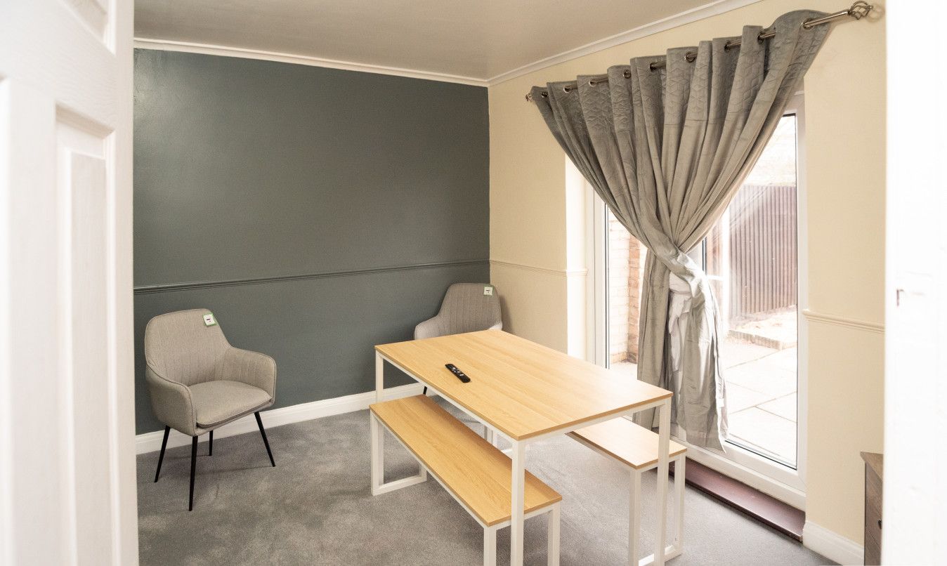 A room with a grey wall, a wooden table, two chairs, a bench, and a window with grey curtains.