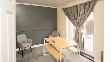 A room with a grey wall, a wooden table, two chairs, a bench, and a window with grey curtains.
